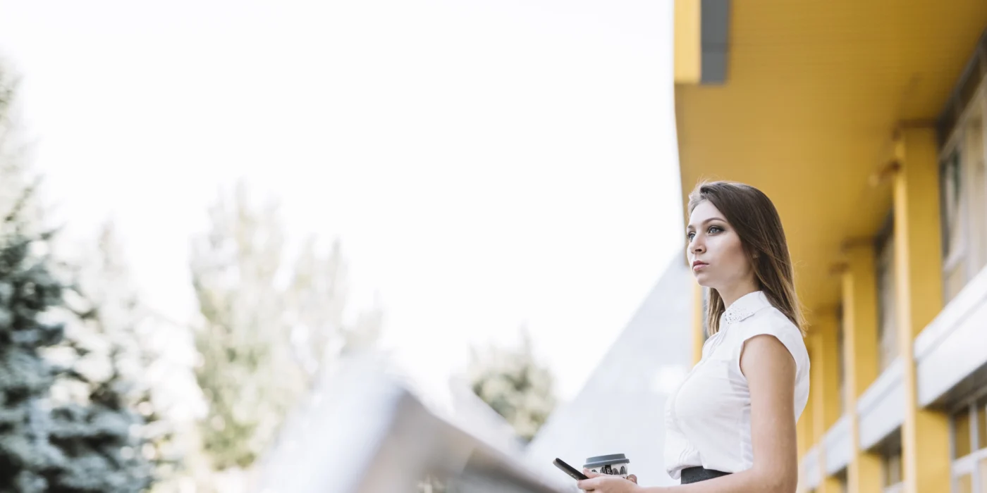 young-businesswoman-standing-balcony-holding-cellphone-disposable-coffee-cup-scaled