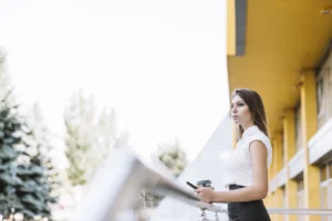 young-businesswoman-standing-balcony-holding-cellphone-disposable-coffee-cup-scaled