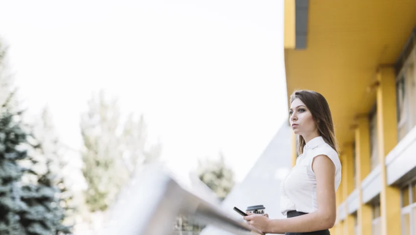 young-businesswoman-standing-balcony-holding-cellphone-disposable-coffee-cup-scaled